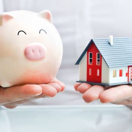 2 hands, one holding a piggy bank, one holding a house model