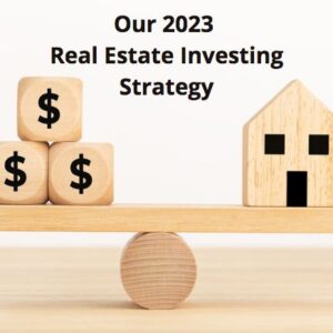 Wooden scale with money blocks on one end and a wooden house on the other. Text title sayind - Our 2023 Real Estate Investing Strategy.